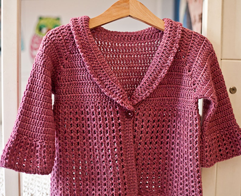May include: A pink crocheted cardigan with a wide collar and button closure. The cardigan has a delicate openwork pattern.