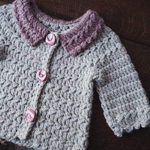 May include: A light gray, hand-crocheted cardigan with a contrasting mauve collar and three pink rose-shaped buttons. The textured stitchwork and scalloped edges add detail to this handmade garment. A cozy, handmade baby or toddler sweater.