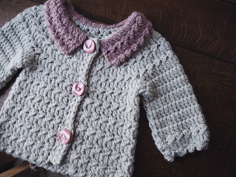 May include: A light gray, hand-crocheted cardigan with a contrasting mauve collar and three pink rose-shaped buttons. The textured stitchwork and scalloped edges add detail to this handmade garment. A cozy, handmade baby or toddler sweater.