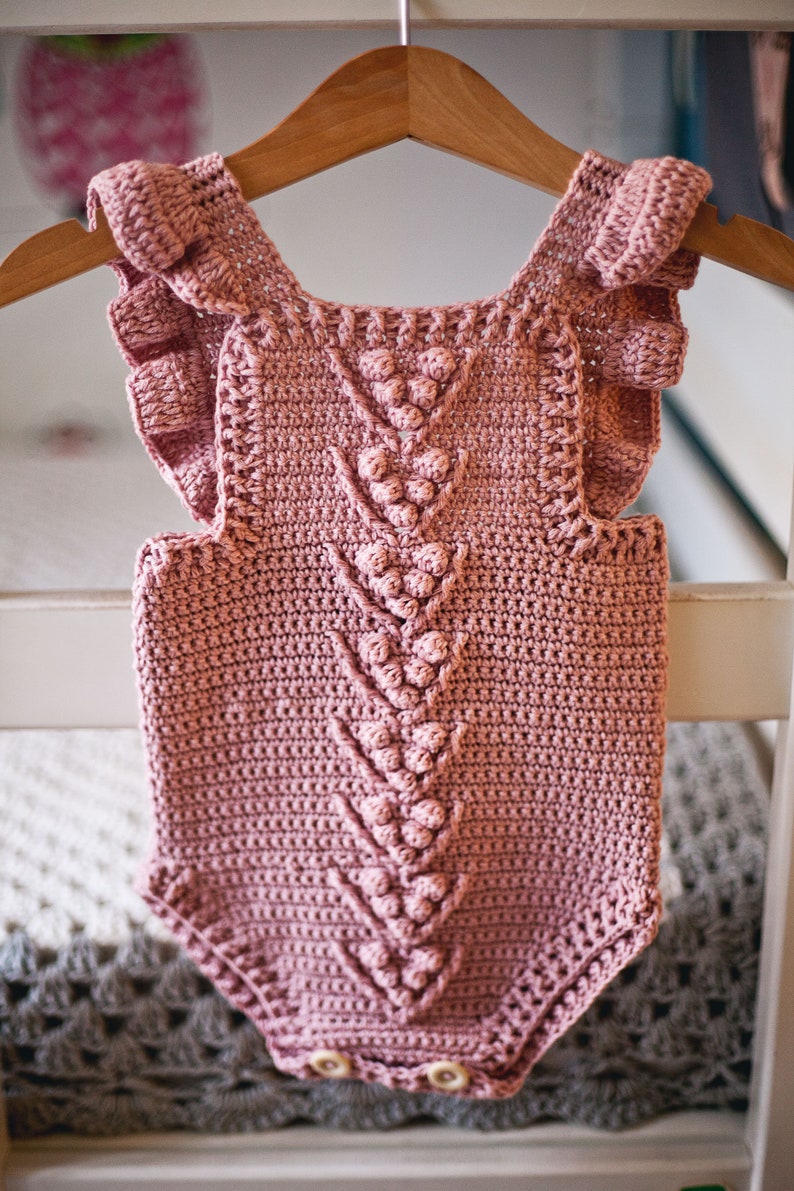 May include: A pink crocheted baby onesie with a ruffled neckline and a decorative pattern of crocheted balls. The onesie has two white buttons at the bottom.