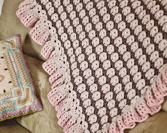 May include: A crocheted baby blanket with a pink and brown diamond pattern and a pink ruffled border.