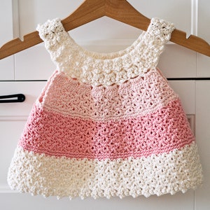 May include: A pink and white crocheted baby dress with a scalloped edge.
