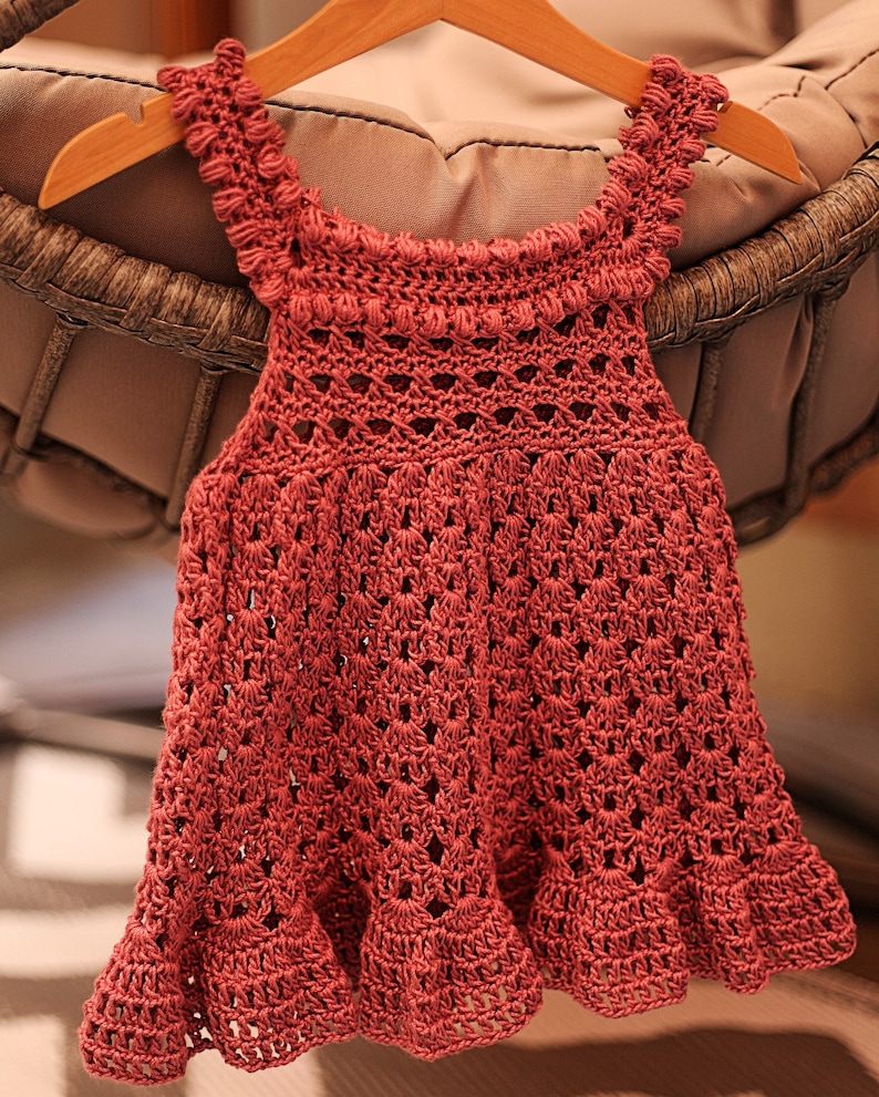 May include: A crocheted baby dress in a deep pink color with a scalloped hem and a delicate lace pattern.