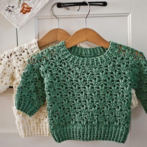 May include: Two crocheted sweaters hanging on wooden hangers. The sweater on the left is cream colored and the sweater on the right is green.