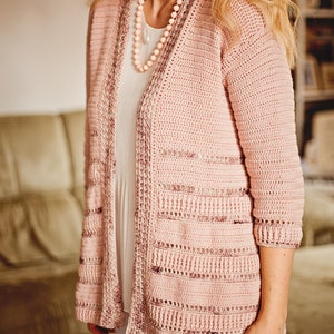 Peut inclure: Un cardigan en crochet rose avec un motif ajour&eacute; d&eacute;licat. Le cardigan est long, ample et &agrave; manches trois-quarts.
