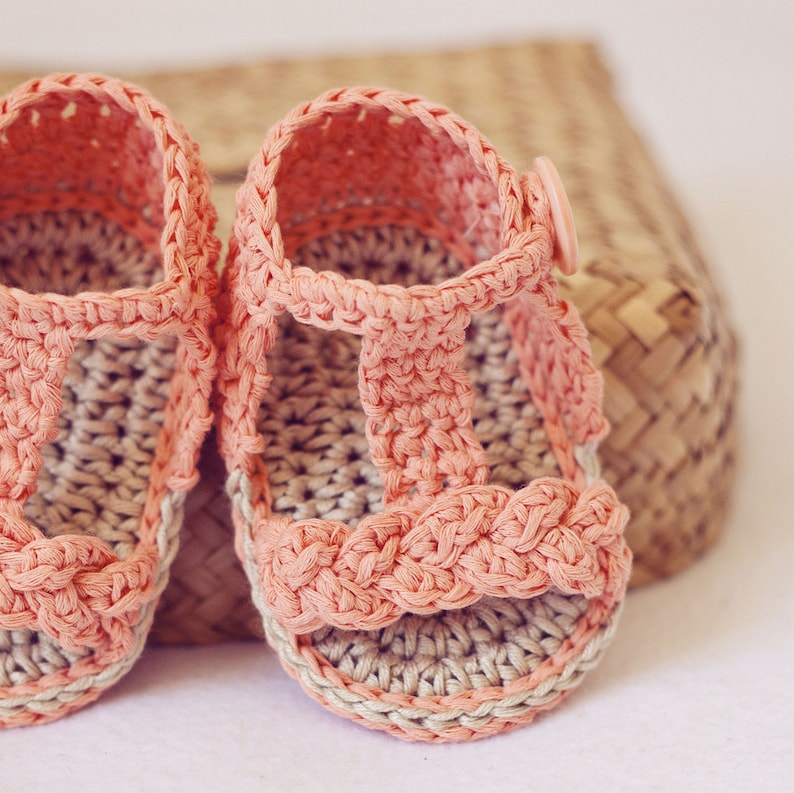 Puede incluir: Un par de sandalias de beb&eacute; de ganchillo en color rosa claro. Las sandalias tienen un dise&ntilde;o de correa en T y est&aacute;n hechas con un hilo beige.