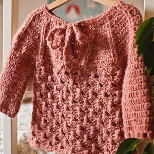 May include: A pink crocheted sweater with a tie at the back. The sweater has a textured pattern and is hanging on a hanger.