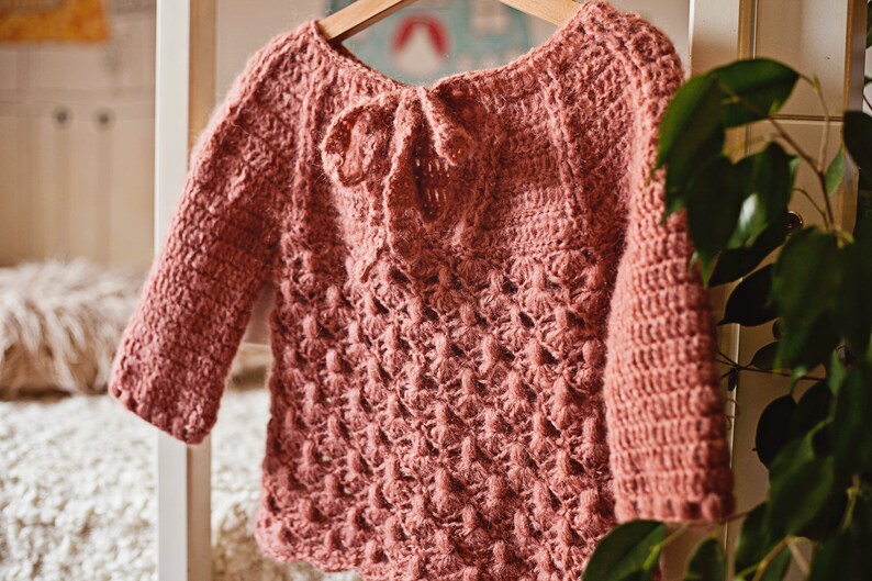 May include: A pink crocheted sweater with a tie at the back. The sweater has a textured pattern and is hanging on a hanger.