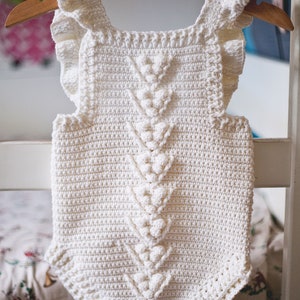 May include: White crocheted baby romper with a ruffled neckline and a decorative stitch pattern. The romper has a V-shaped design in the front and is hanging on a wooden hanger.