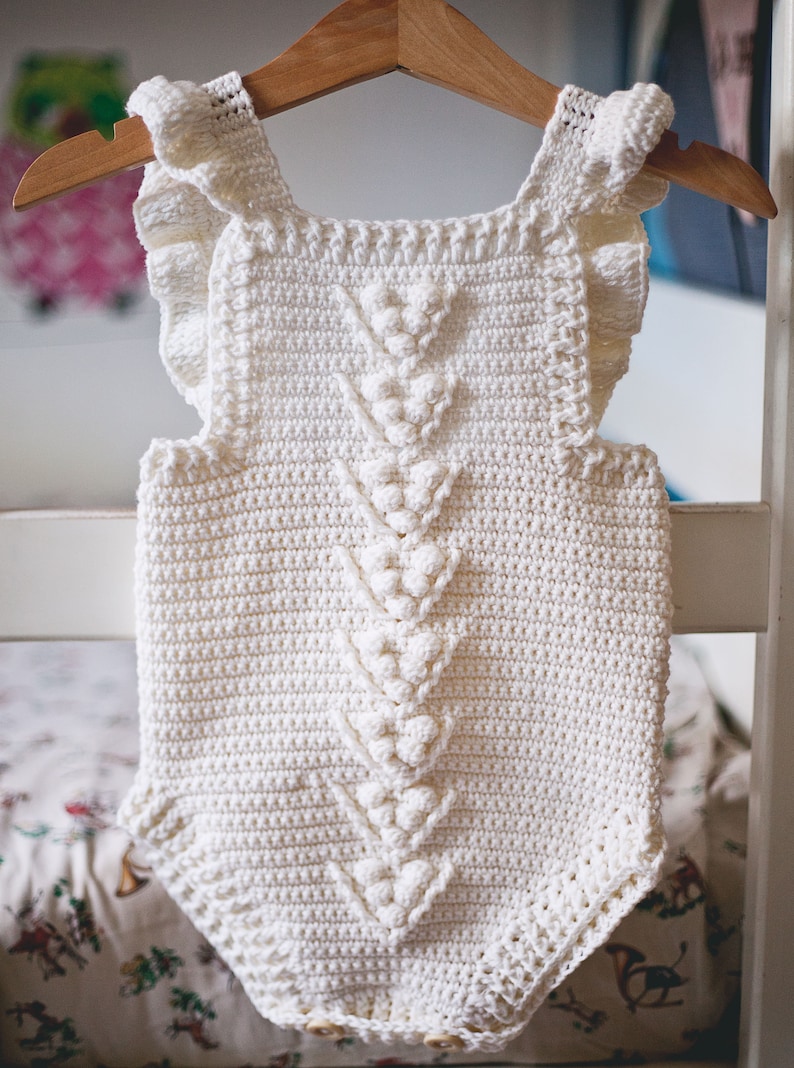 May include: White crocheted baby romper with a ruffled neckline and a decorative stitch pattern. The romper has a V-shaped design in the front and is hanging on a wooden hanger.