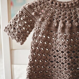 Puede incluir: Un vestido de beb&eacute; de ganchillo marr&oacute;n con un delicado patr&oacute;n de calado y una fila de pompones de ganchillo alrededor del escote.