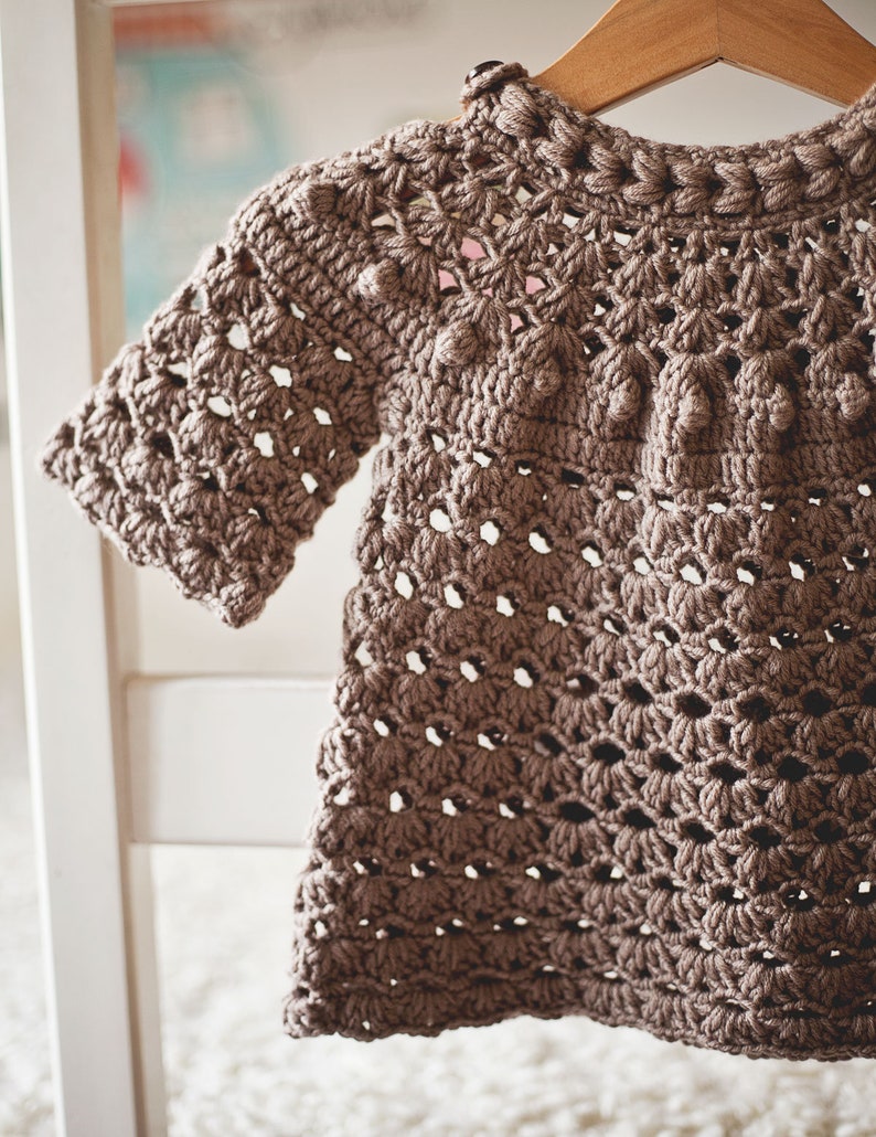Puede incluir: Un vestido de beb&eacute; de ganchillo marr&oacute;n con un delicado patr&oacute;n de calado y una fila de pompones de ganchillo alrededor del escote.