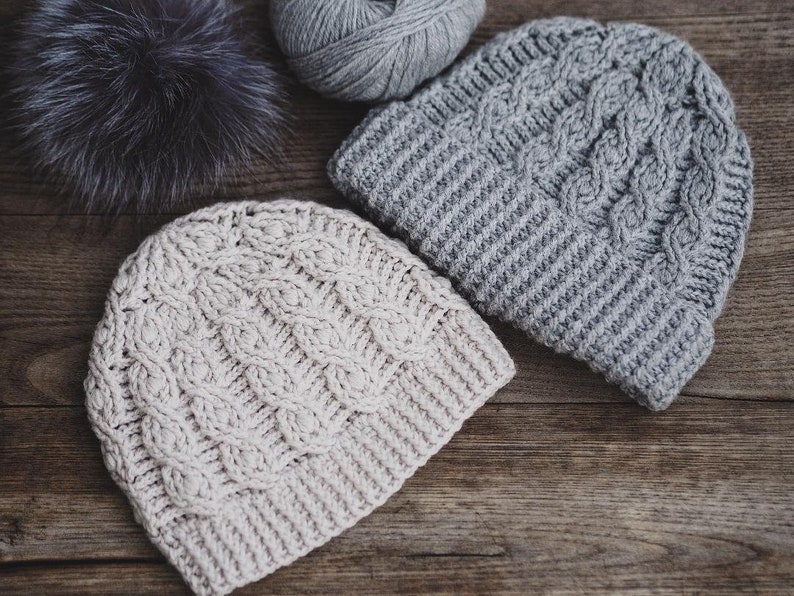 May include: Two crocheted hats, one in beige and one in gray, with a cable knit pattern. The hats are lying on a wooden surface.