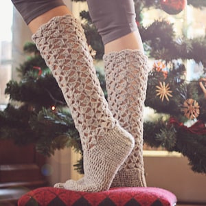 May include: A pair of beige crochet leg warmers with a lacy pattern. The leg warmers are worn by a person standing in front of a Christmas tree.