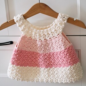 May include: A crocheted baby dress with a pink and white color gradient. The dress has a scalloped neckline and a ruffled hem.