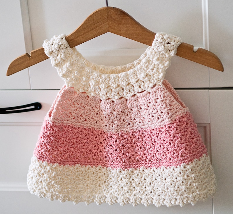 May include: A crocheted baby dress with a pink and white color gradient. The dress has a scalloped neckline and a ruffled hem.
