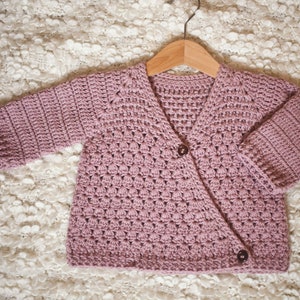 May include: A soft pink crocheted cardigan with a button closure. The cardigan is hanging on a wooden hanger and has a textured stitch pattern.