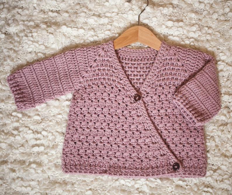 May include: A soft pink crocheted cardigan with a button closure. The cardigan is hanging on a wooden hanger and has a textured stitch pattern.