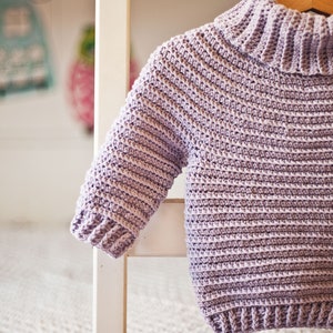 May include: A light purple crocheted sweater with a turtleneck collar. The sweater is hanging on a white wooden chair.