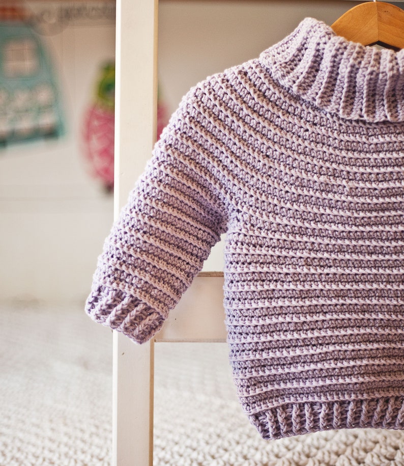 May include: A light purple crocheted sweater with a turtleneck collar. The sweater is hanging on a white wooden chair.