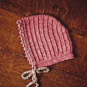 May include: A pink and beige crocheted baby bonnet with a tie. The bonnet has a scalloped edge and is made with a textured stitch.