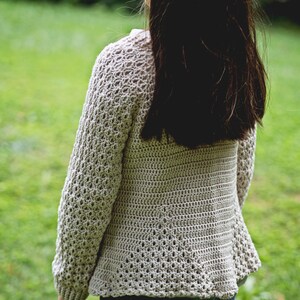 K&ouml;nnte beinhalten: Hellbeige H&auml;kel-Cardigan mit langen &Auml;rmeln und einem strukturierten Lochmuster. Der Cardigan wird &uuml;ber einer blauen Jeans getragen. Das Bild ist im Freien mit einem gr&uuml;nen, grasbewachsenen Hintergrund aufgenommen.