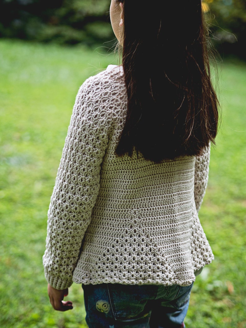 K&ouml;nnte beinhalten: Hellbeige H&auml;kel-Cardigan mit langen &Auml;rmeln und einem strukturierten Lochmuster. Der Cardigan wird &uuml;ber einer blauen Jeans getragen. Das Bild ist im Freien mit einem gr&uuml;nen, grasbewachsenen Hintergrund aufgenommen.