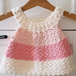 May include: A crocheted baby dress with a pink and white ombre design. The dress has a scalloped hem and a crocheted collar.