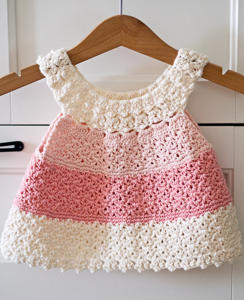 May include: A crocheted baby dress with a pink and white ombre design. The dress has a scalloped hem and a crocheted collar.