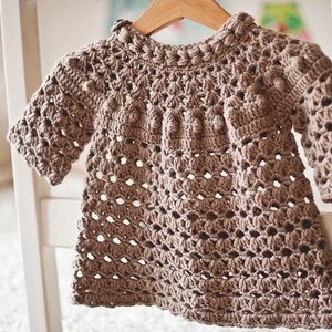 Puede incluir: Un jersey de beb&eacute; de color marr&oacute;n tejido a crochet con un delicado patr&oacute;n de calado y un escote redondo. El jersey tiene una forma ligeramente acampanada y es perfecto para una ocasi&oacute;n especial.