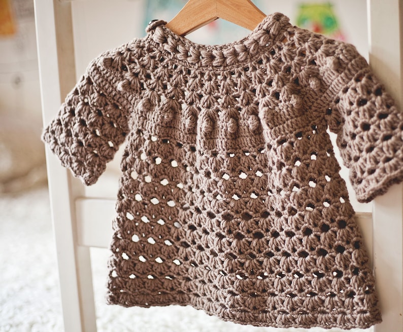 Puede incluir: Un jersey de beb&eacute; de color marr&oacute;n tejido a crochet con un delicado patr&oacute;n de calado y un escote redondo. El jersey tiene una forma ligeramente acampanada y es perfecto para una ocasi&oacute;n especial.