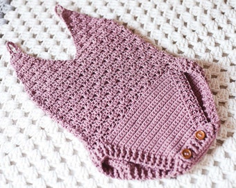 May include: A crocheted baby onesie in a dusty rose color with a delicate openwork pattern. The onesie has a sleeveless design and features two wooden buttons on the bottom.