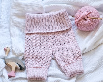 Puede incluir: Pantalones de bebé de crochet rosa con una cintura doblada. Los pantalones están colocados sobre un fondo de tela blanca con un ovillo de hilo rosa y una aguja de tejer de madera.