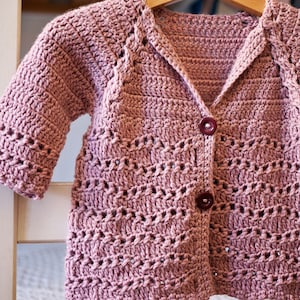 May include: A pink crocheted cardigan with a button closure. The cardigan has a delicate openwork pattern and is hanging on a wooden hanger.