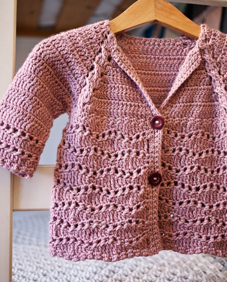 May include: A pink crocheted cardigan with a button closure. The cardigan has a delicate openwork pattern and is hanging on a wooden hanger.