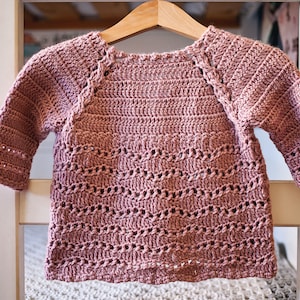 May include: A pink crocheted sweater with a delicate stitch pattern. The sweater has a round neckline and long sleeves.