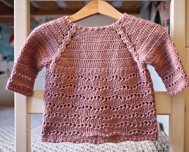 May include: A pink crocheted sweater with a delicate stitch pattern. The sweater has a round neckline and long sleeves.