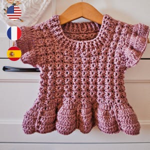 May include: A dusty rose crochet top with a scalloped hem and short sleeves. The handmade garment is displayed on a wooden hanger. The top features a textured stitch pattern and a round neckline.