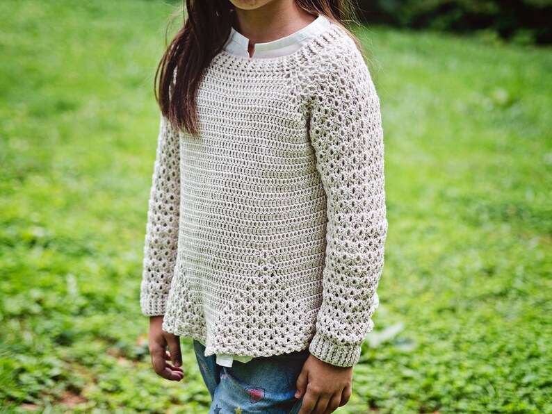 K&ouml;nnte beinhalten: Ein hellbeige H&auml;kelpullover mit langen &Auml;rmeln. Der Pullover hat einen Rundhalsausschnitt und einen leicht ausgestellten Saum. Die &Auml;rmel haben ein offeneres, dekoratives Stichmuster. Der Pullover wird &uuml;ber einem wei&szlig;en Hemd getragen.