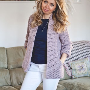 May include: A light purple crocheted cardigan with a shawl collar and pockets. The cardigan is worn over a navy blue t-shirt and white pants.