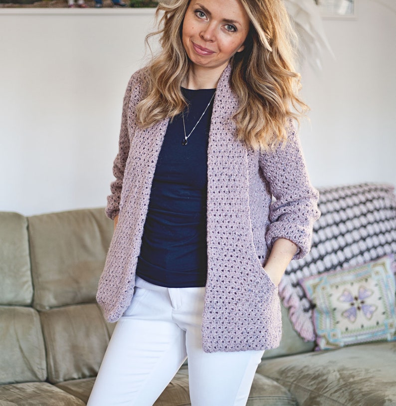 May include: A light purple crocheted cardigan with a shawl collar and pockets. The cardigan is worn over a navy blue t-shirt and white pants.