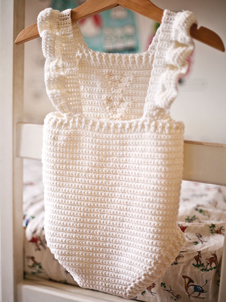 May include: White crocheted baby romper with ruffled straps. The romper is hanging on a wooden hanger.