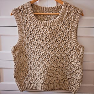 Könnte beinhalten: Beige Häkelweste mit rundem Ausschnitt und ärmellosem Design. Die Weste ist aus einem strukturierten, offen gewebten Stich gefertigt und hängt an einem Holzbügel. Das Kleidungsstück hat eine neutrale Farbe und eignet sich zum Tragen in Schichten.