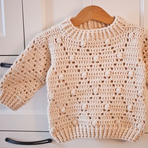 May include: A cream-colored crocheted sweater with a textured stitch pattern and a round neckline. The sweater has a loose fit and is perfect for layering.