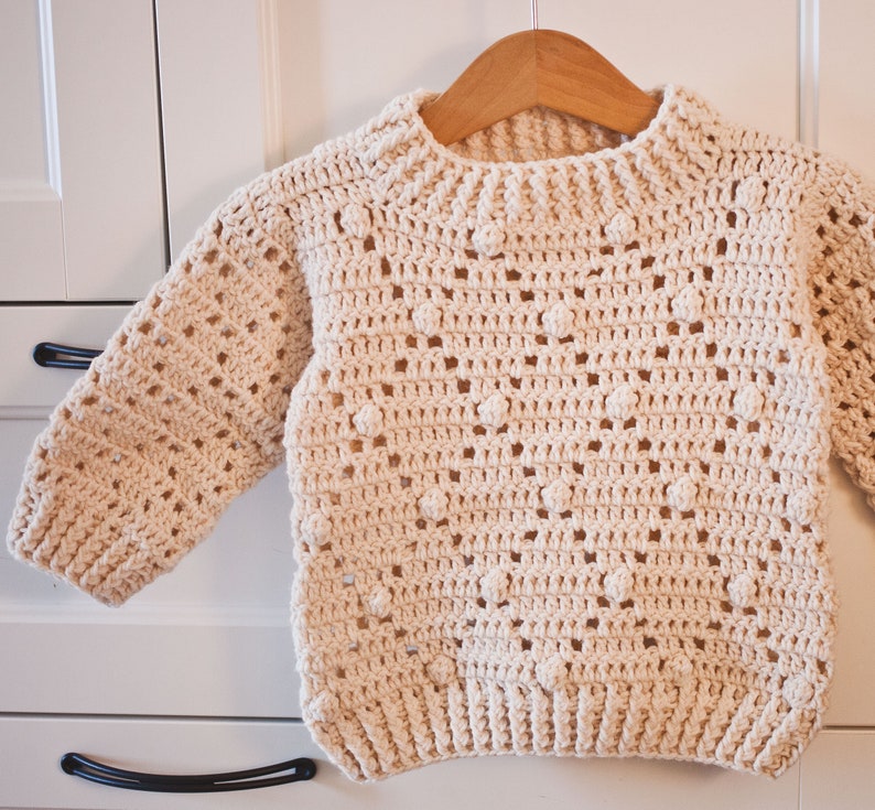 May include: A cream-colored crocheted sweater with a textured stitch pattern and a round neckline. The sweater has a loose fit and is perfect for layering.
