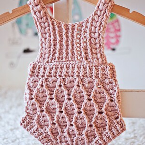 Op de afbeelding: Een roze gebreide baby romper met een gegolfde zoom en een delicaat patroon. De romper hangt aan een houten hanger.