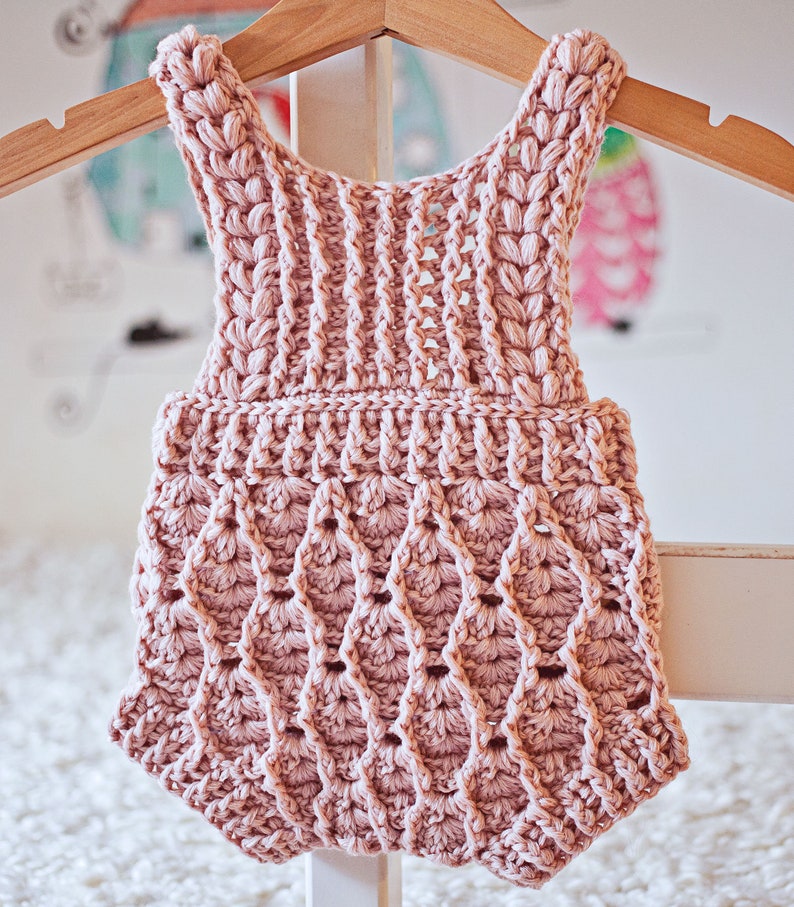 Op de afbeelding: Een roze gebreide baby romper met een gegolfde zoom en een delicaat patroon. De romper hangt aan een houten hanger.