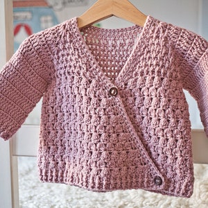 May include: A pink crocheted cardigan with a button closure. The cardigan has a textured stitch pattern and is hanging on a white hanger.