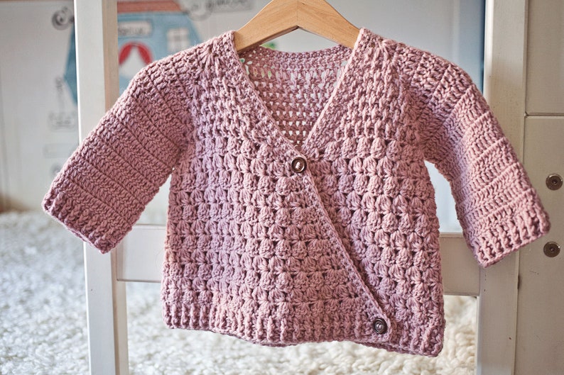 May include: A pink crocheted cardigan with a button closure. The cardigan has a textured stitch pattern and is hanging on a white hanger.