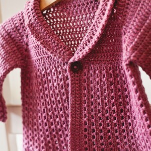 May include: A pink crocheted cardigan with a button closure and a wide collar. The cardigan is hanging on a wooden hanger.